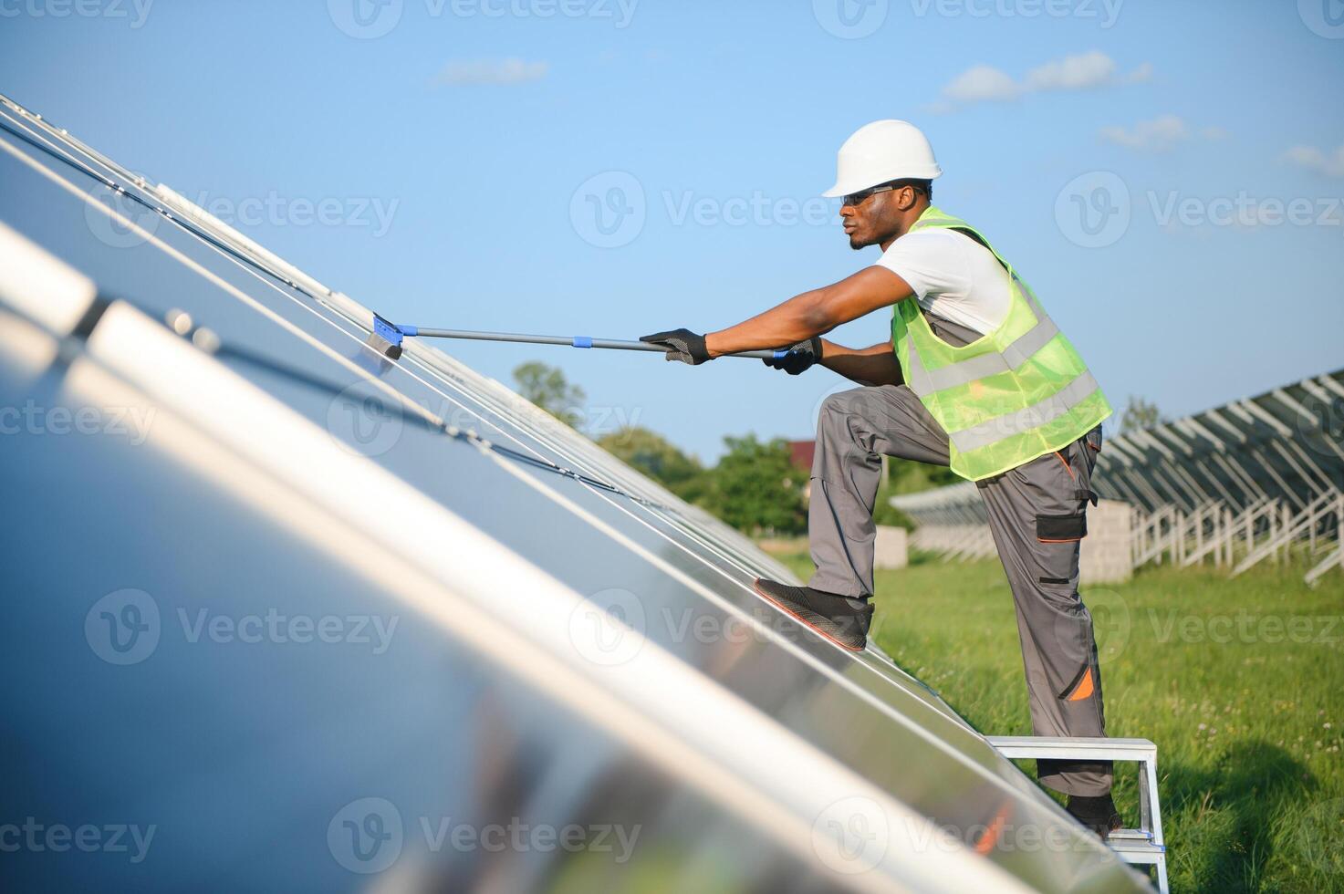 Alternative power plant worker in uniform cleaning solar panels with mop. Handsome African-American taking care of equipment. photo