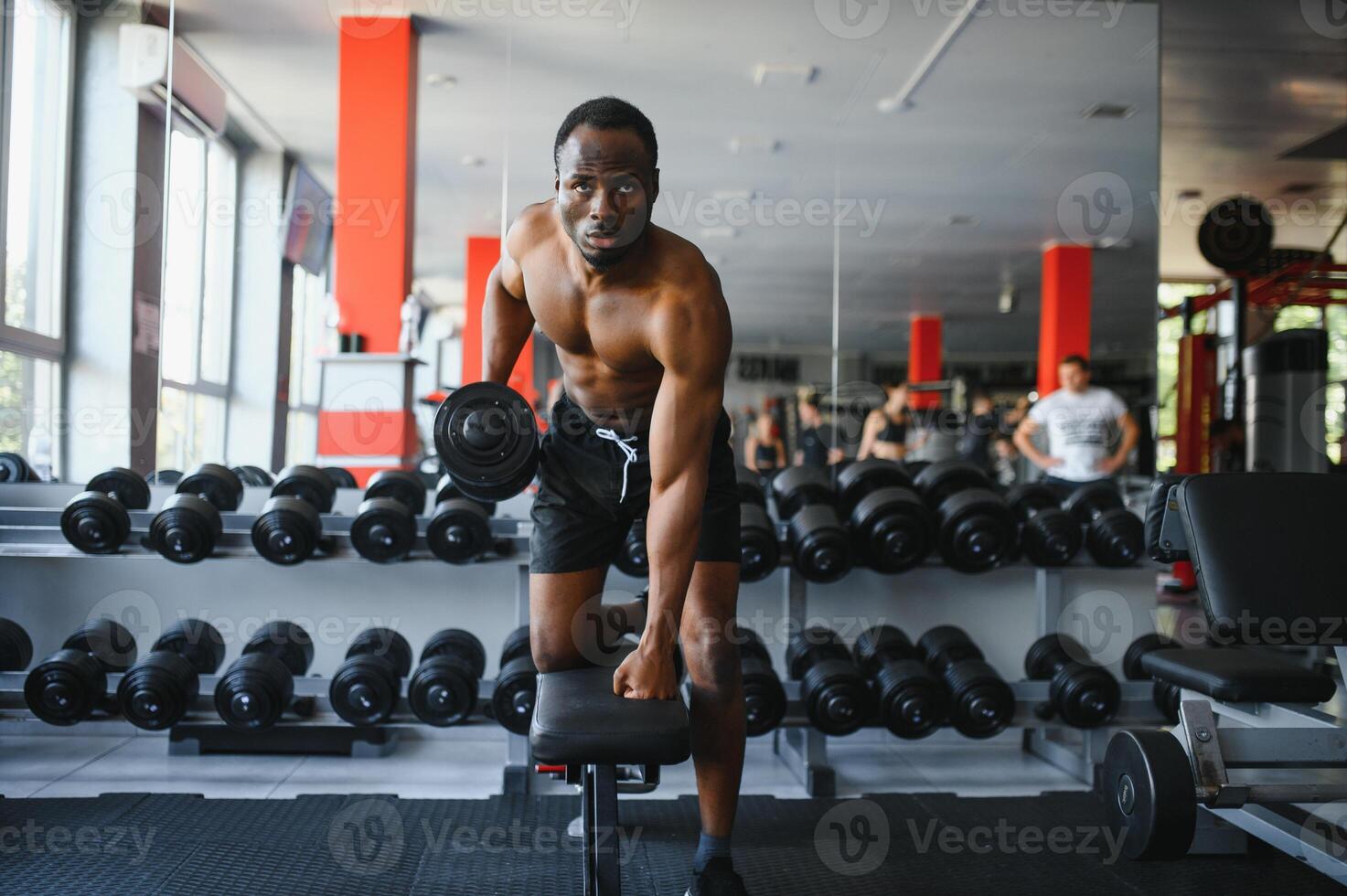 Young African American man sitting and lifting a dumbbell close to the rack at gym. Male weight training person doing a biceps curl in fitness center photo