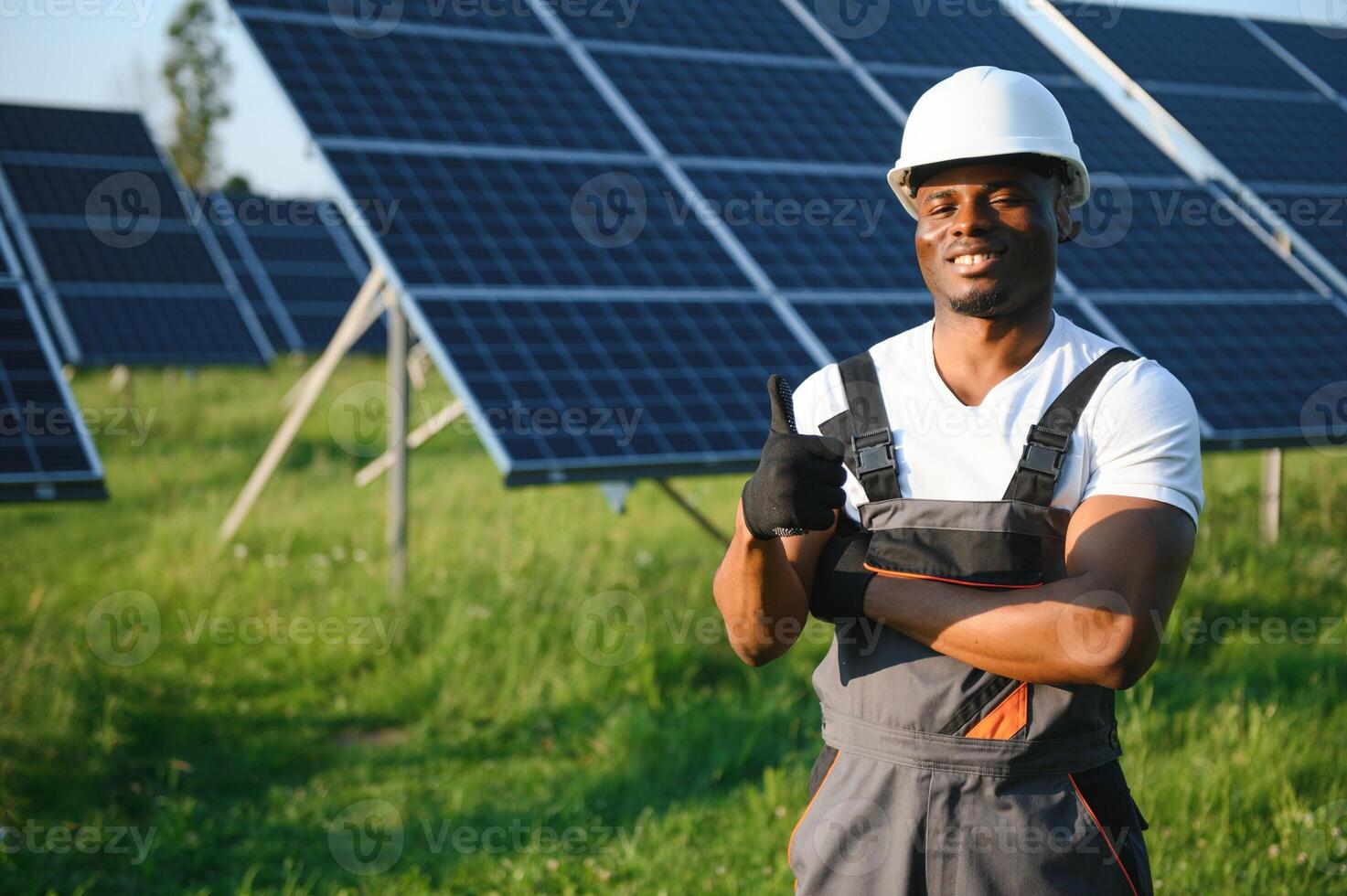 retrato de joven hermoso africano americano artesano en protector casco. hombre en uniforme y con herramientas en pie entre solar paneles foto