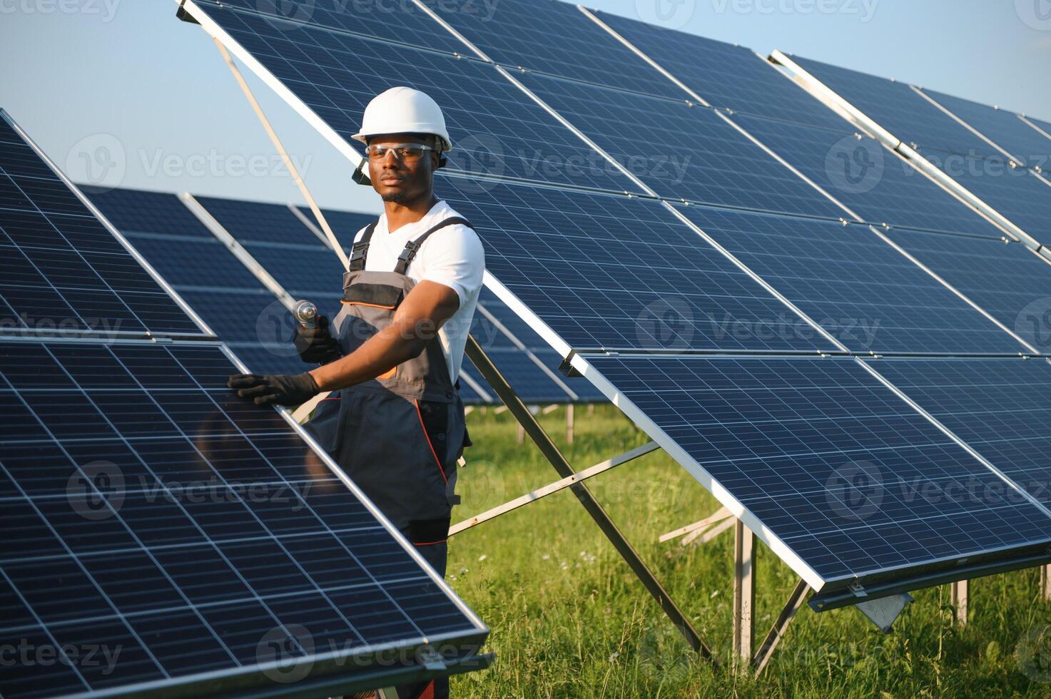 africano americano hombre en la seguridad casco y lentes apretar nueces en solar paneles con destornillador. competente técnico utilizando herramientas mientras ejecutando Servicio trabajo en estación foto