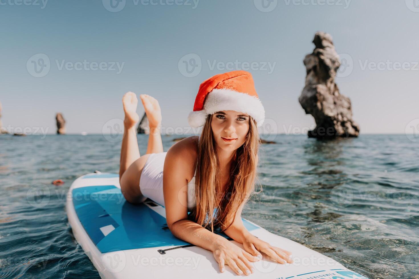 Woman sea sup. Close up portrait of happy young caucasian woman with long hair in Santa hat looking at camera and smiling. Cute woman portrait in a white bikini posing on sup board in the sea photo