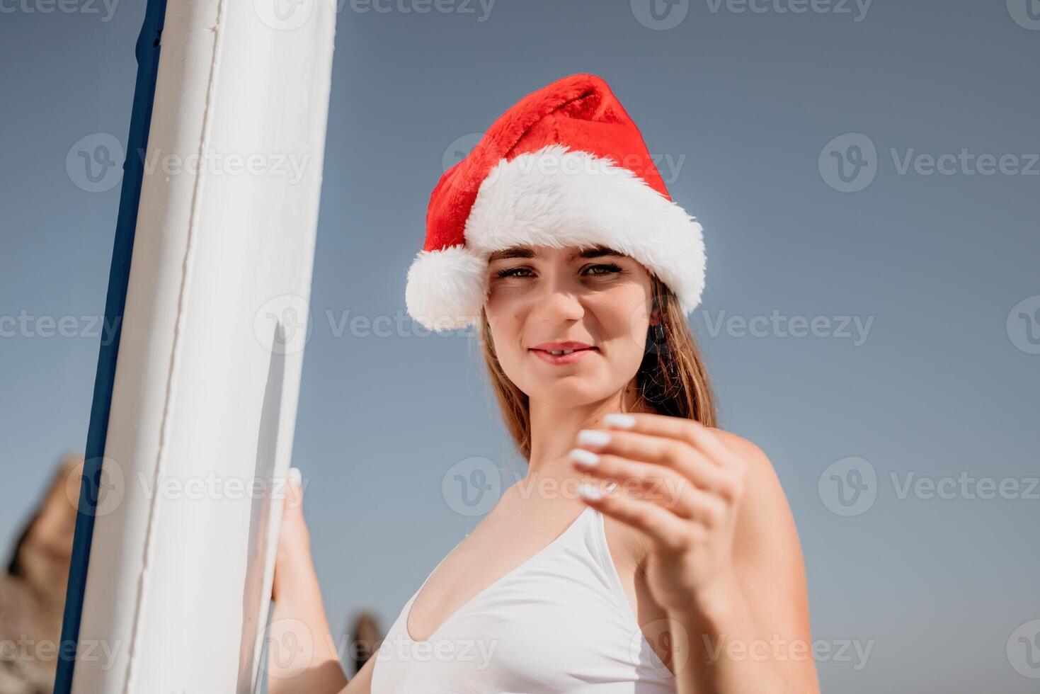 mujer mar sorber. cerca arriba retrato de contento joven caucásico mujer con largo pelo en Papa Noel sombrero mirando a cámara y sonriente. linda mujer retrato en un blanco bikini posando en cenar tablero en el mar foto