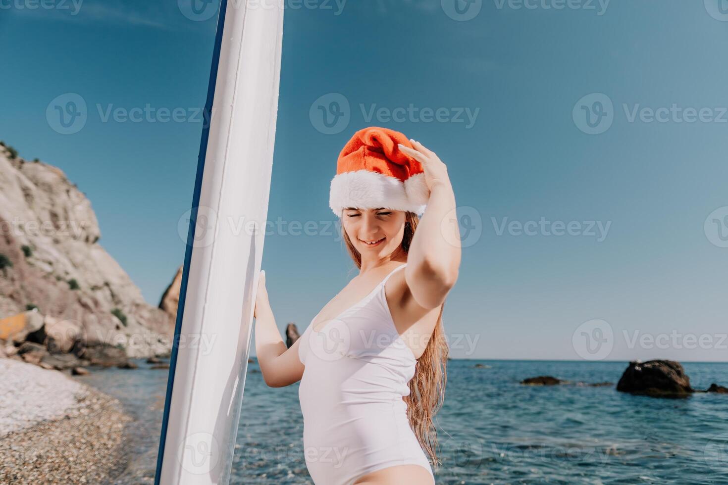 Woman sea sup. Close up portrait of happy young caucasian woman with long hair in Santa hat looking at camera and smiling. Cute woman portrait in a white bikini posing on sup board in the sea photo