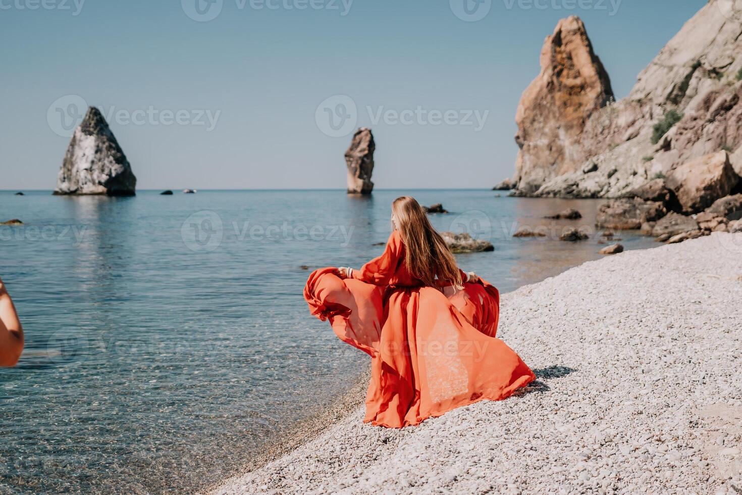 Woman travel sea. Happy tourist in red dress enjoy taking picture outdoors for memories. Woman traveler posing on the rock at sea bay surrounded by volcanic mountains, sharing travel adventure journey photo