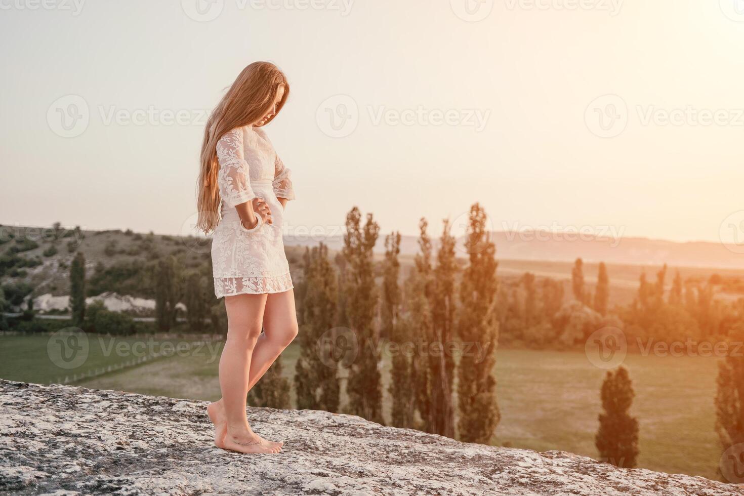 Happy woman in white boho dress on sunset in mountains. Romantic woman with long hair standing with her back on the sunset in nature in summer with open hands. Silhouette. Nature. Sunset. photo