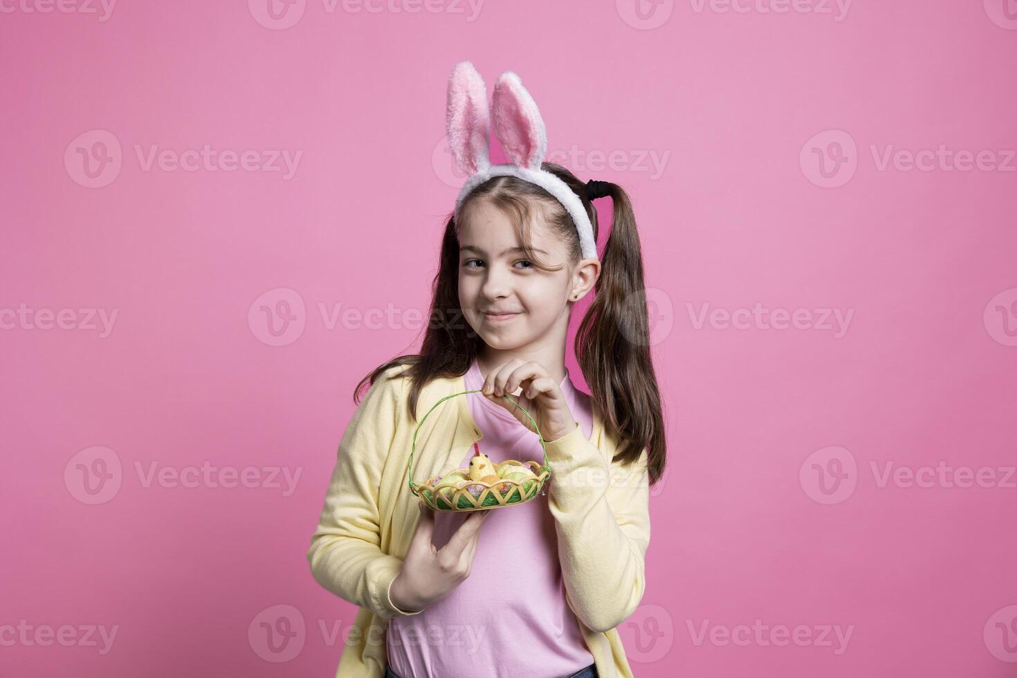 Young joyful child posing with easter eggs and toys on camera, feeling happy and excited about spring holiday celebration and gifts. Little girl with bunny ears smiling in studio. photo