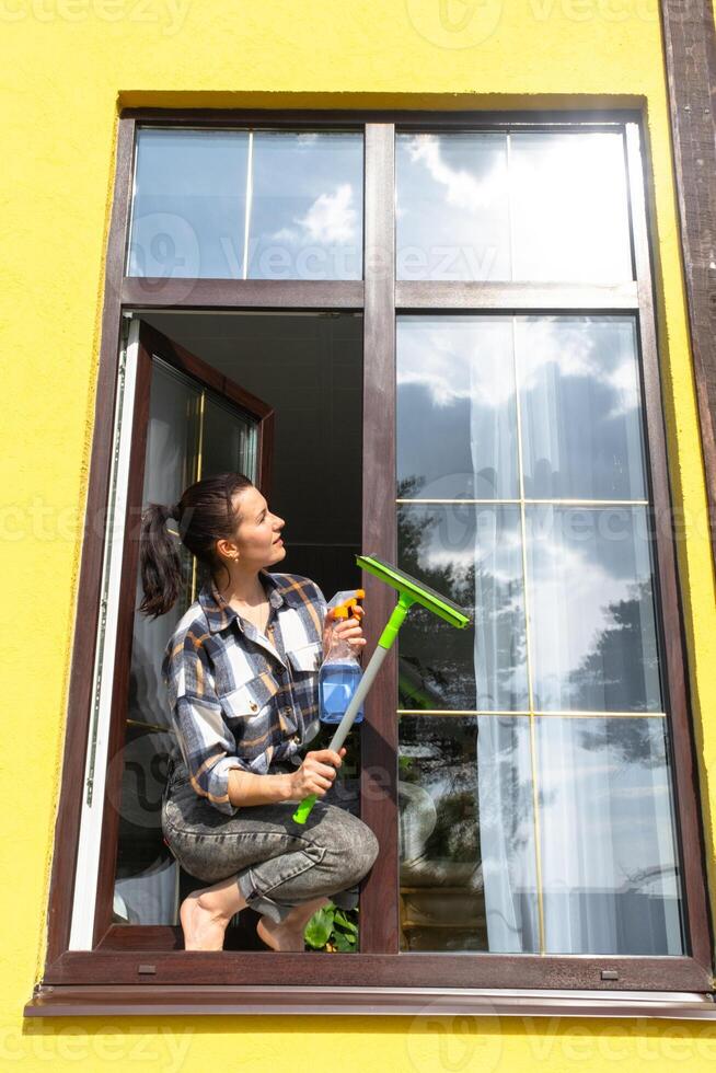 un mujer a mano lavados el ventana de el casa con un trapo con un rociar limpiador y un fregona afuera. la seguridad a altura, restaurar orden y limpieza en el primavera, limpieza Servicio foto