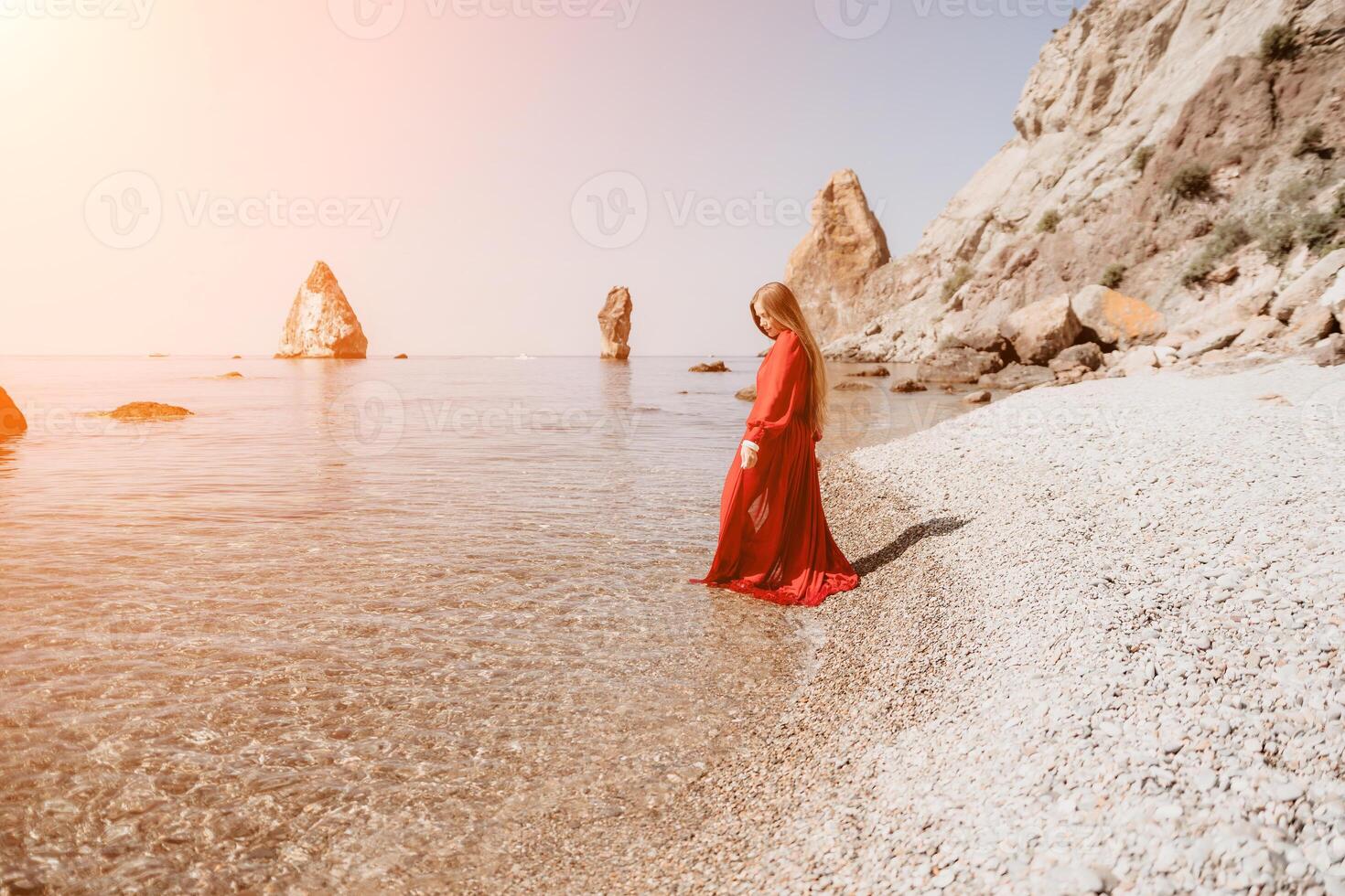 Woman travel sea. Happy tourist in red dress enjoy taking picture outdoors for memories. Woman traveler posing on the rock at sea bay surrounded by volcanic mountains, sharing travel adventure journey photo