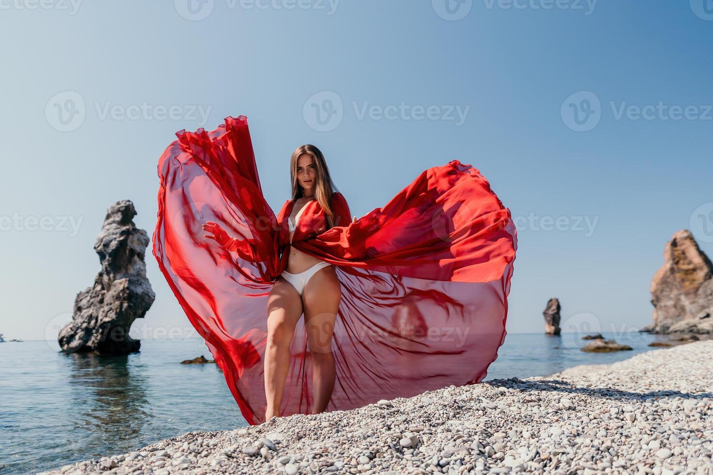 Woman travel sea. Happy tourist in red dress enjoy taking picture outdoors for memories. Woman traveler posing on the rock at sea bay surrounded by volcanic mountains, sharing travel adventure journey photo