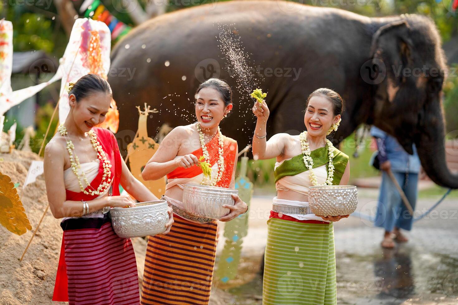 Closeup beautiful Thai young lady ware Thai traditional dress use flowers to sprinkle water on each other on the Thai New Year's Day in a fun way on blurred elephant and pile of sand background. photo