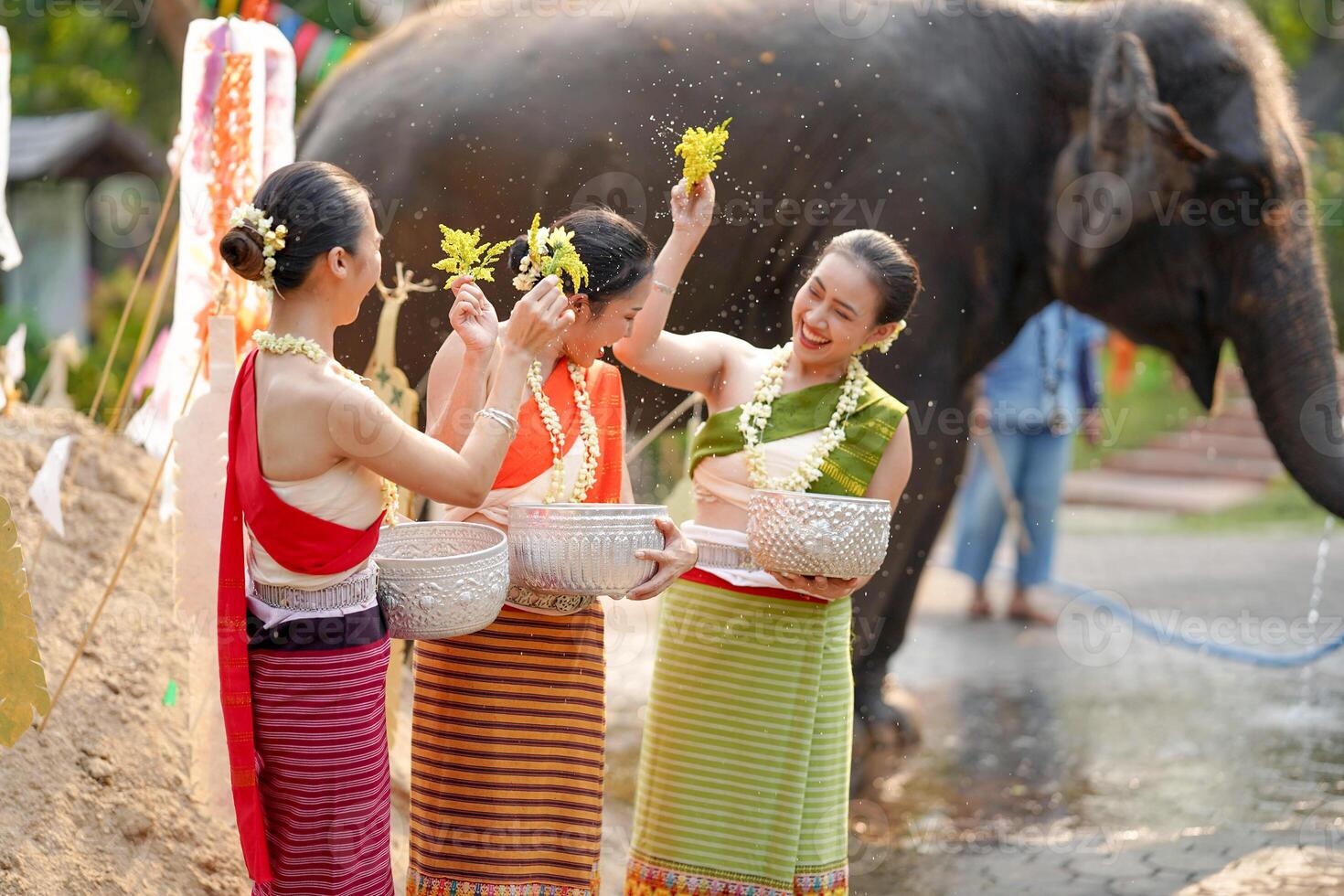 Beautiful Thai young lady ware Thai traditional dress use flowers to sprinkle water on each other on the Thai New Year's Day in a fun way on blurred elephant and pile of sand background. photo