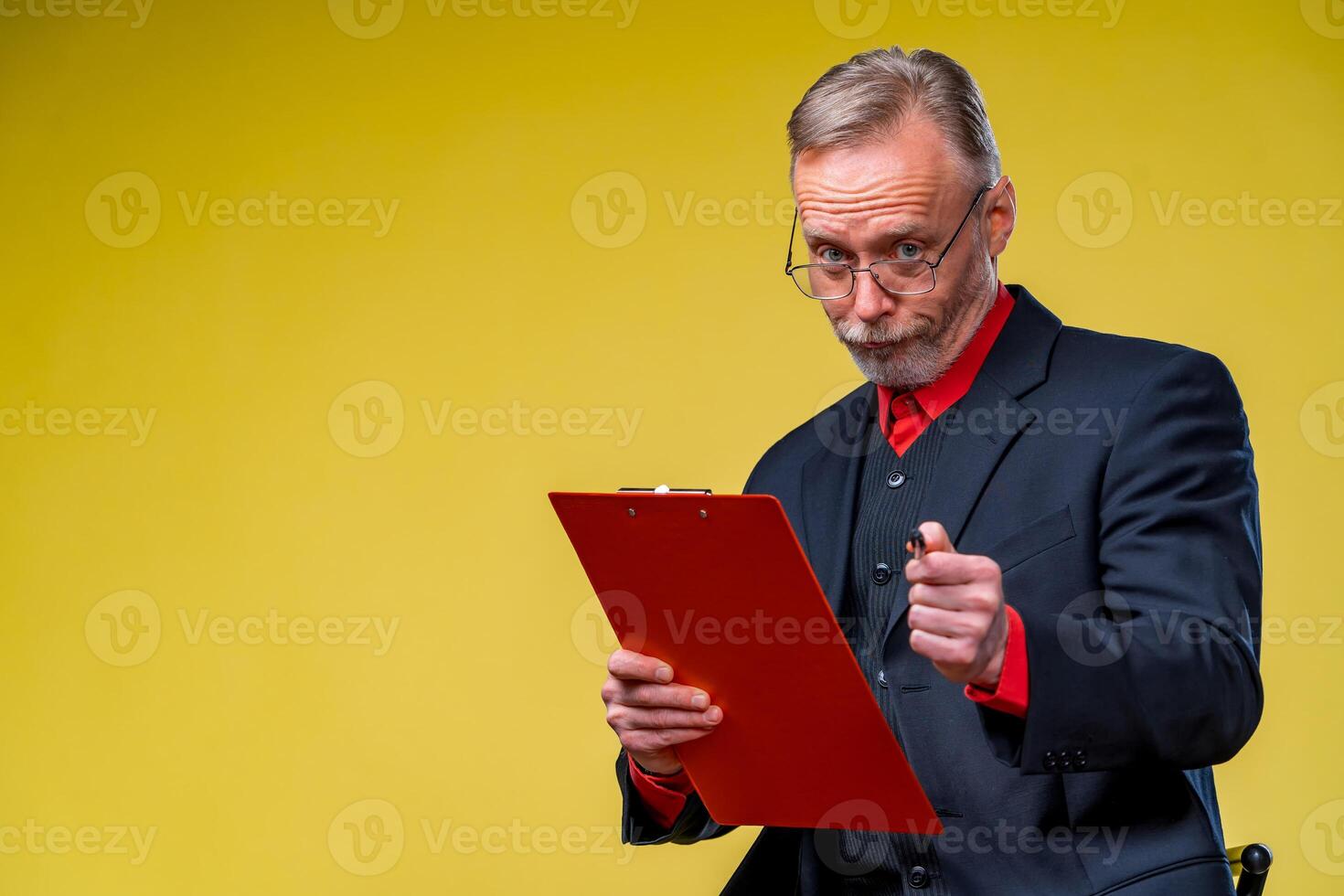 Senior businessman sitting with folder over yellow background in suit looking at camera in red shirt . Business concept photo