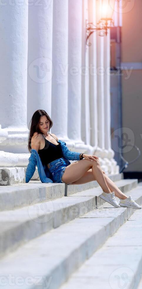 Fashionable brunette young woman posing on steps. White columns background. Jeans shirt and shorts. photo