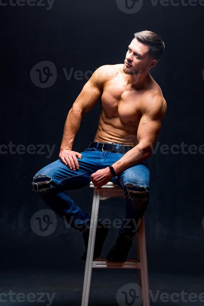 Man with perfect body sitting on high chair. Dark background. Male beauty. photo