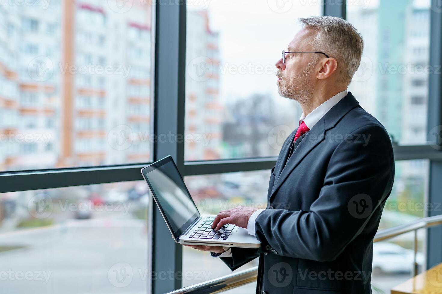 Man looking away from window. Laptop in hands, man thinking while checking e-mails standing near window. Photo from the side