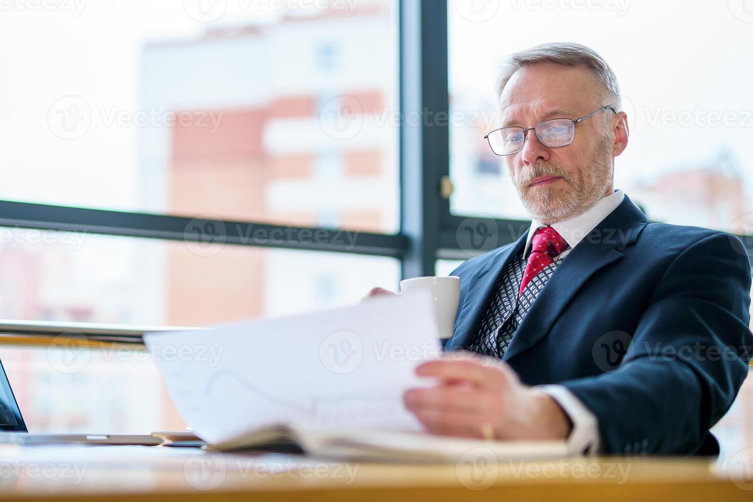 Businessman is checking a printed report or document while sitting at table, working on paper work at a laptop explaining his point of view. Photo in an office.