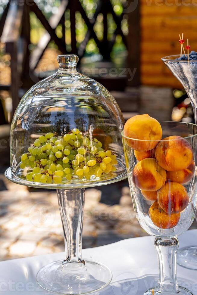 Table decoration with glass and crystal tableware. Grapes under dome cover and peaches in vase. Closeup. photo