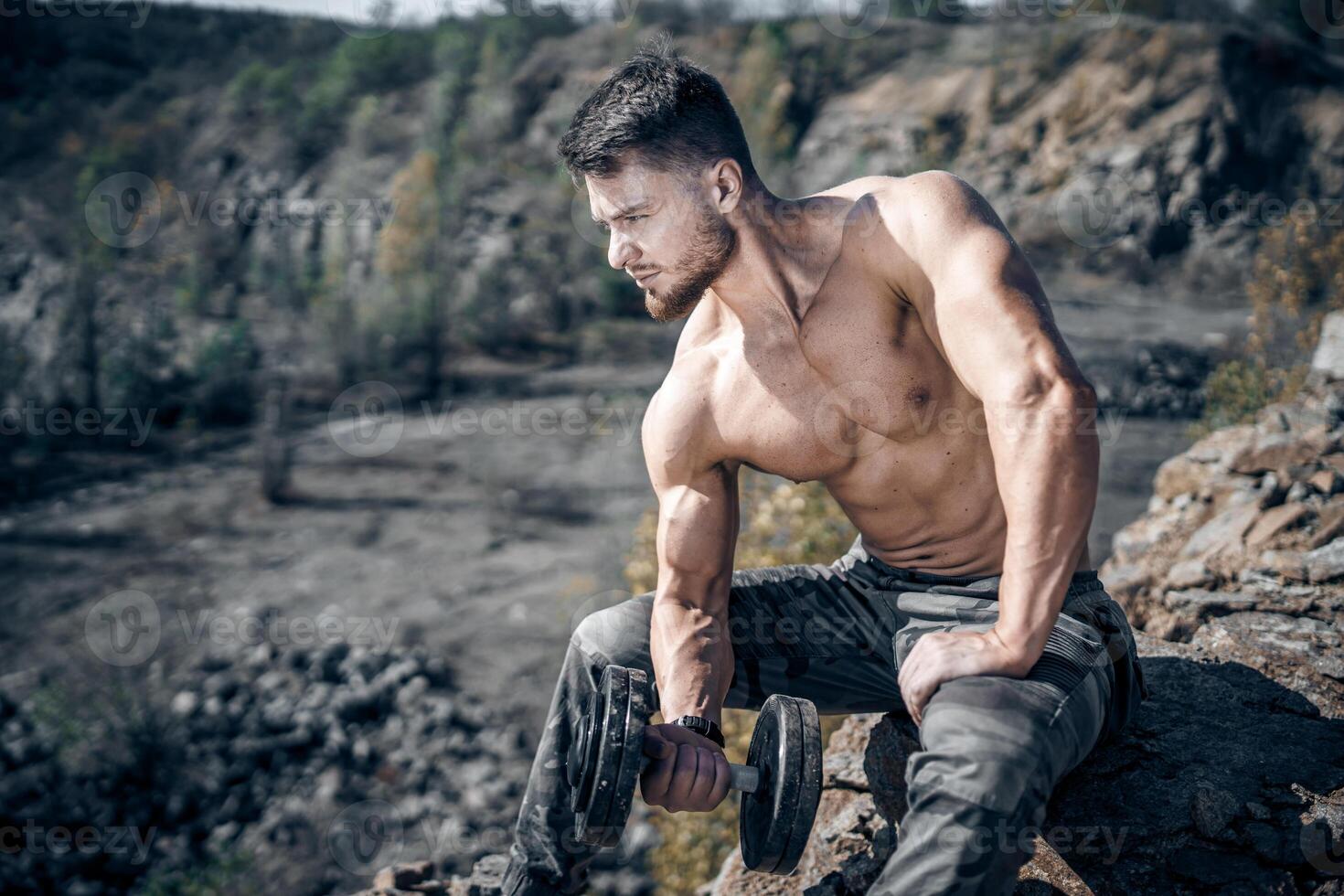 Athletic man sitting on a rock with dumbbell. Quarry background. Outdoor training. photo