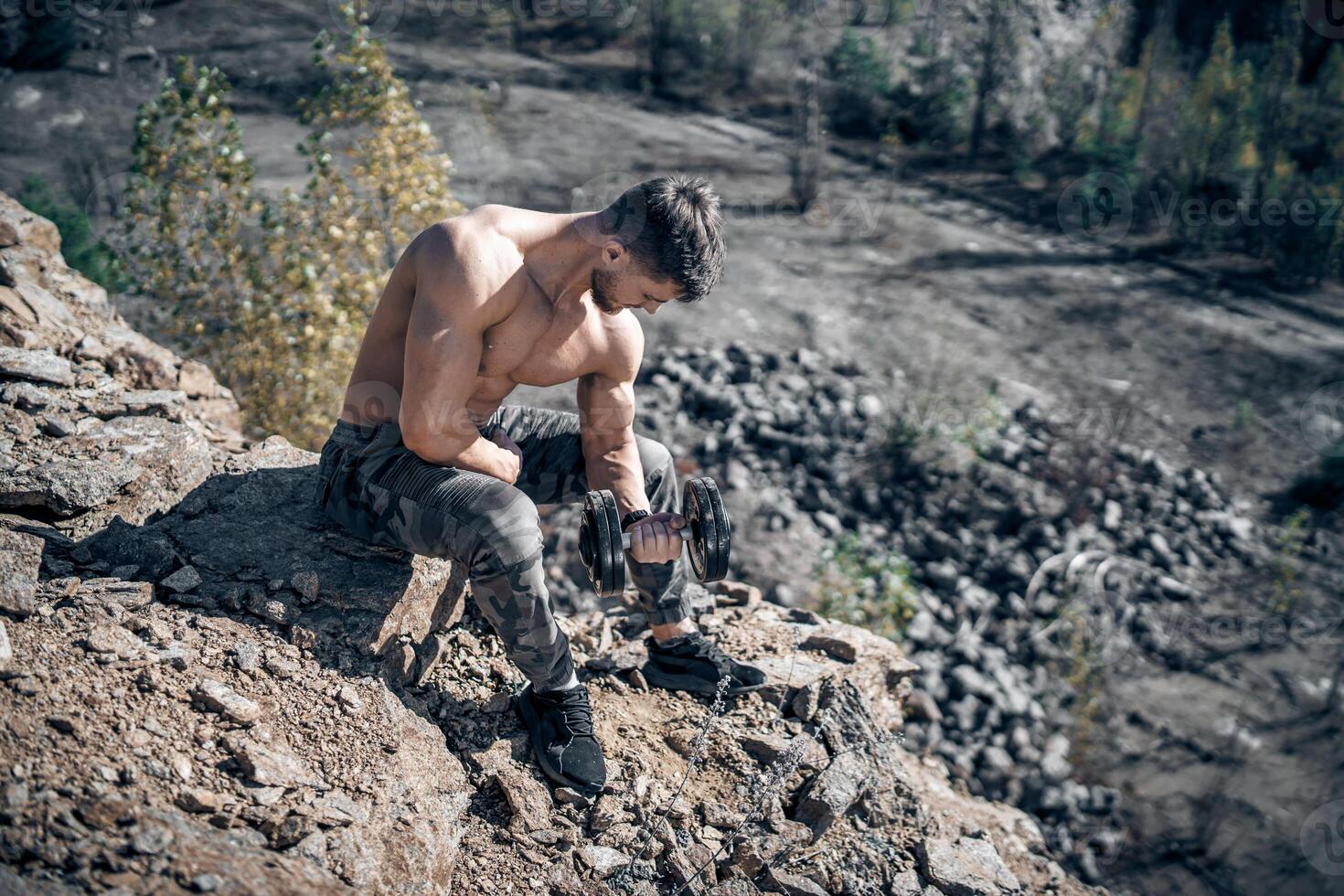 Full size photo of an athletic guy posing with rock in a quarry. Outdoor shot. Bodybuilder. Quarry background. Closeup. Dumbbell in hand.