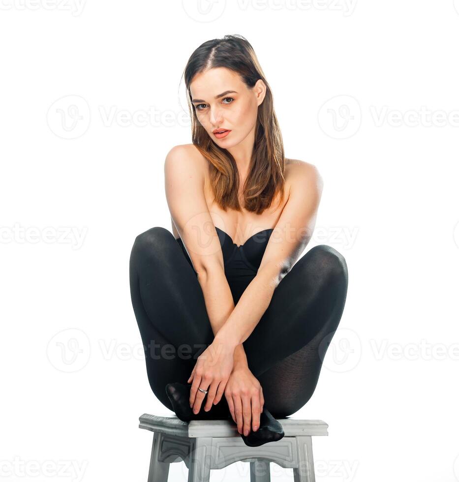 Pretty woman posing sitting in black top and leggings on a chair over white background. Woman with long dark hair sits with crossed hands and crossed legs. photo