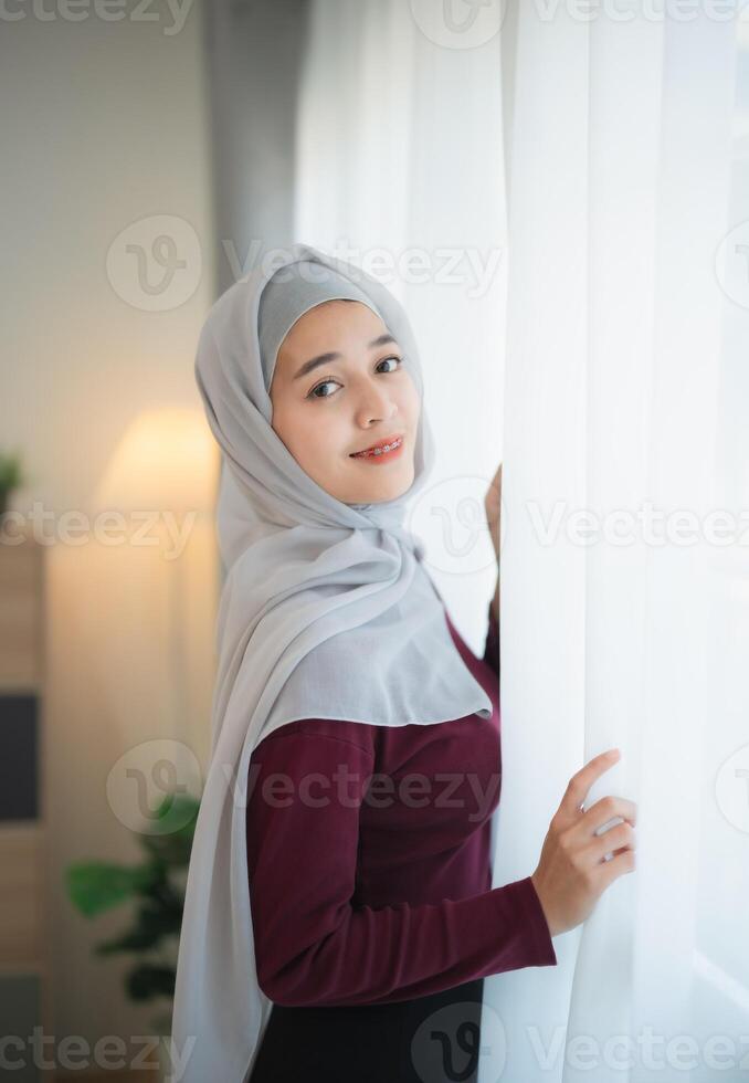 Muslim Islam woman wearing hijab or grey scarf and a red shirt is standing in front of a window. She is smiling and looking at the camera photo