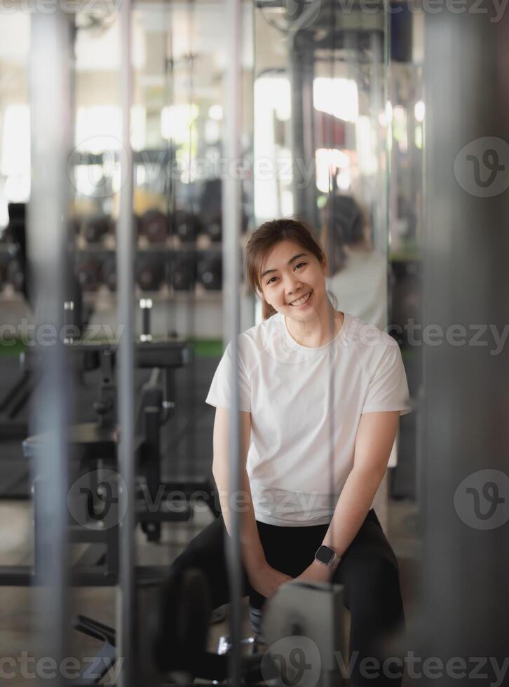 asiático deporte mujer es sentado en un peso máquina con su brazos cruzado a aptitud gimnasia. ella es sonriente y mirando a el cámara. aptitud gimnasio deporte concepto. foto