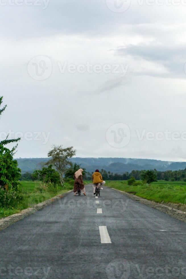 dos hijab asiático mujer montando bicicleta a el Mañana en el asfalto la carretera. dos muchachas ciclismo con montaña y arrozal arroz campo ver foto