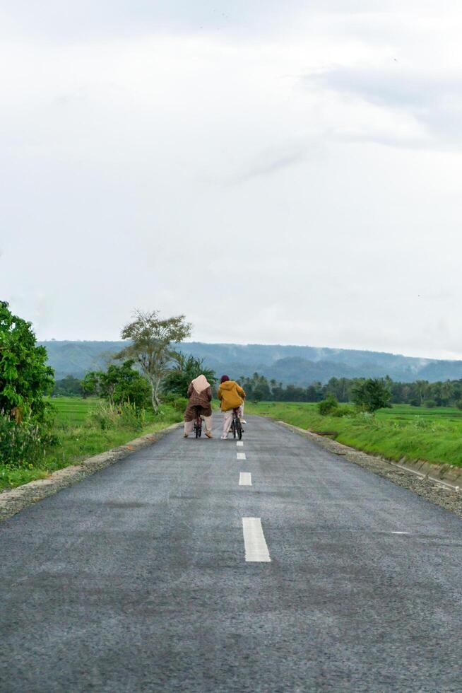 Two hijab Asian woman riding bicycle at the morning on the asphalt road. Two girls cycling with mountain and paddy rice field view photo