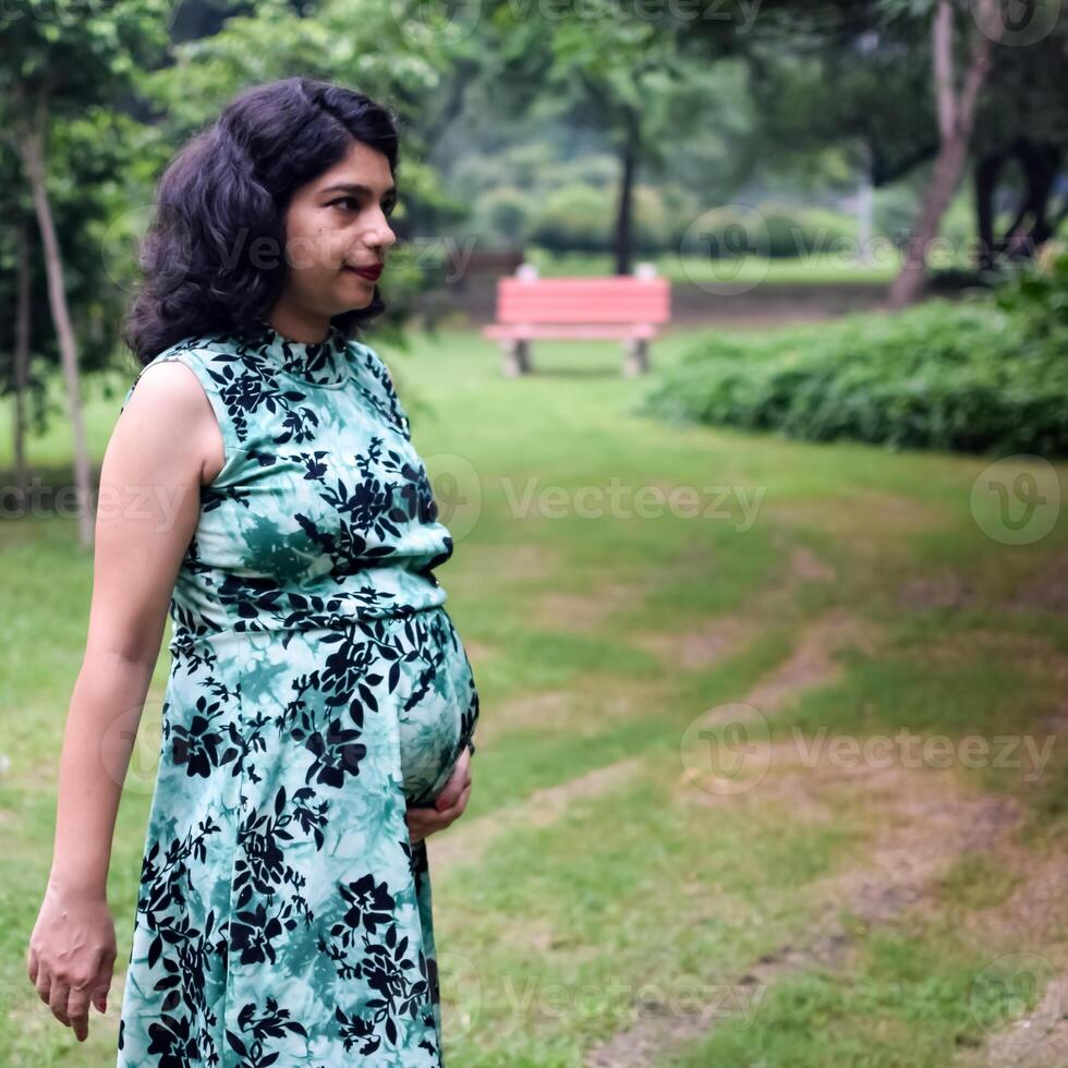 A pregnant Indian lady poses for outdoor pregnancy shoot and hands on belly, Indian pregnant woman puts her hand on her stomach with a maternity dress at society park, Pregnant outside maternity shoot photo