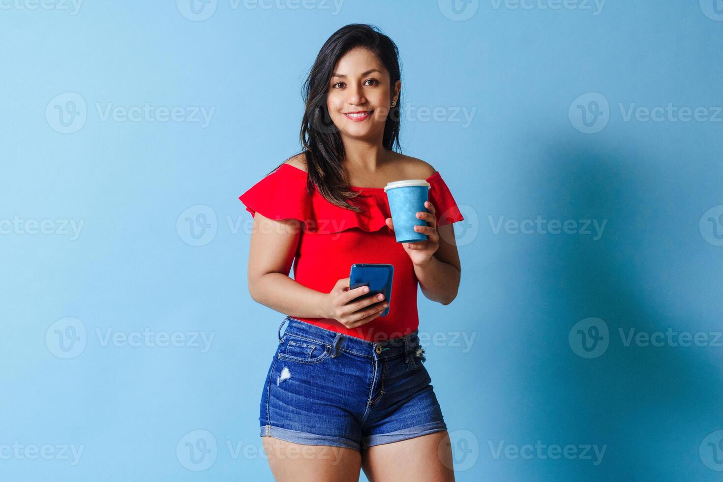 portrait young latinx woman using mobile phone holding takeaway coffee cup look at camera. photo