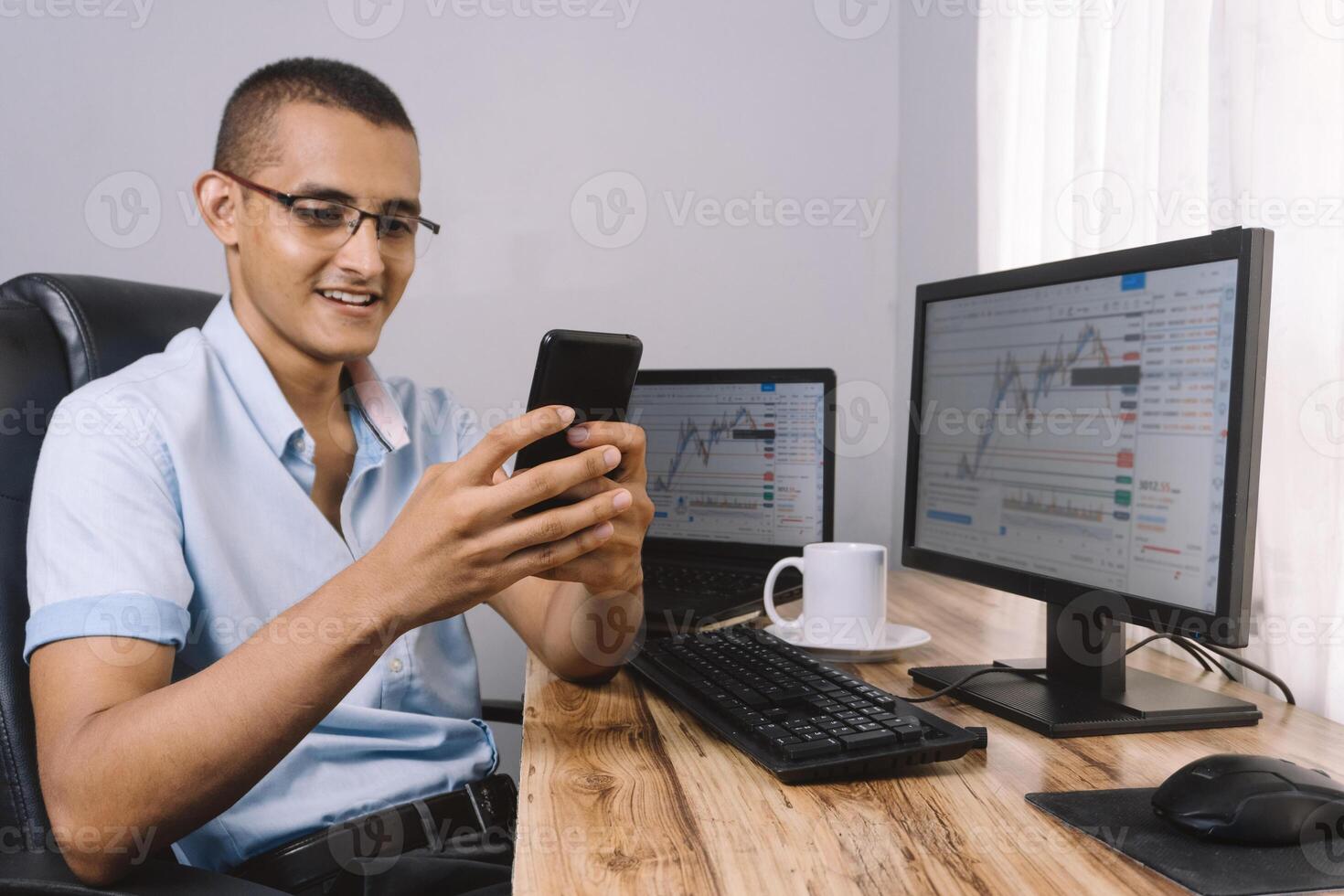 young Hispanic man using his mobile phone in front of his computer. portrait of young investor and cheerful trader watching his smartphone. photo