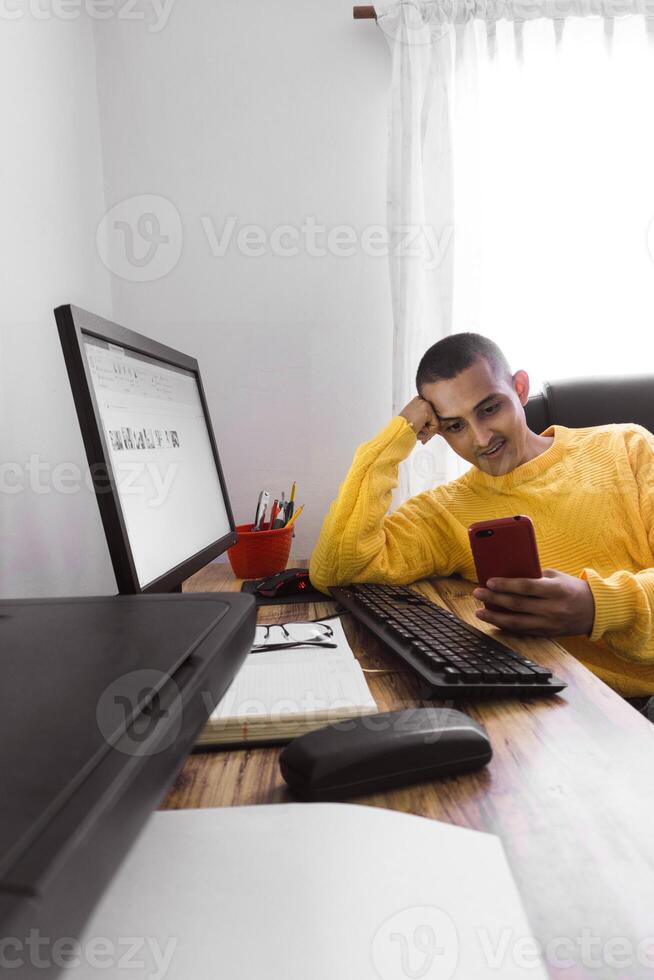 cheerful young hispanic man looking at a phone sitting at a desk indoor. photo