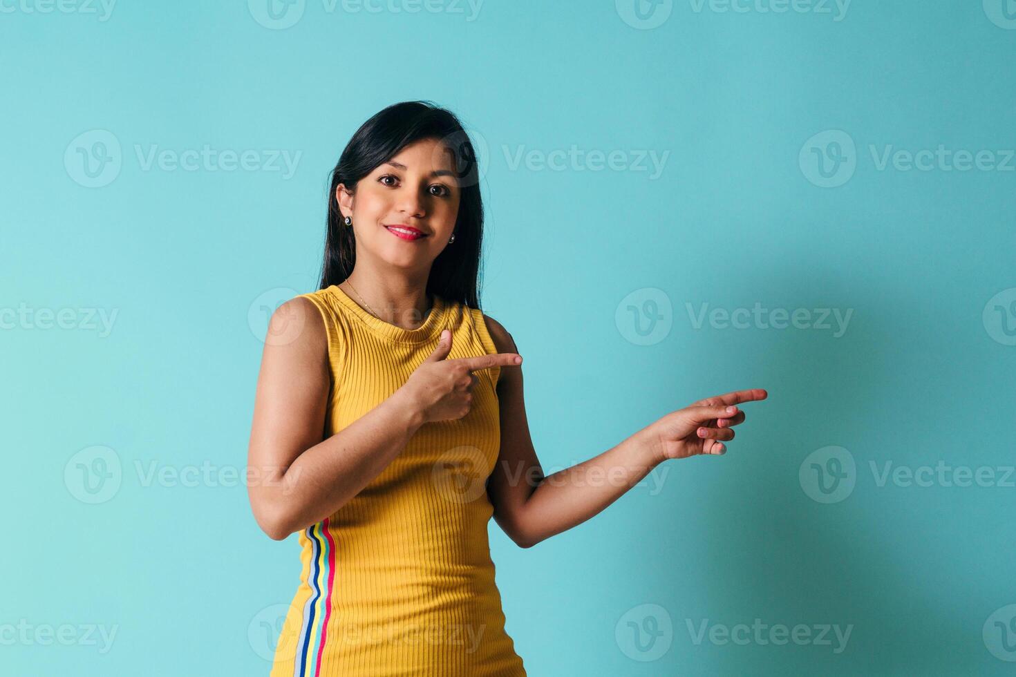 young latin american woman pointing away and looking at the camera over blue background. photo