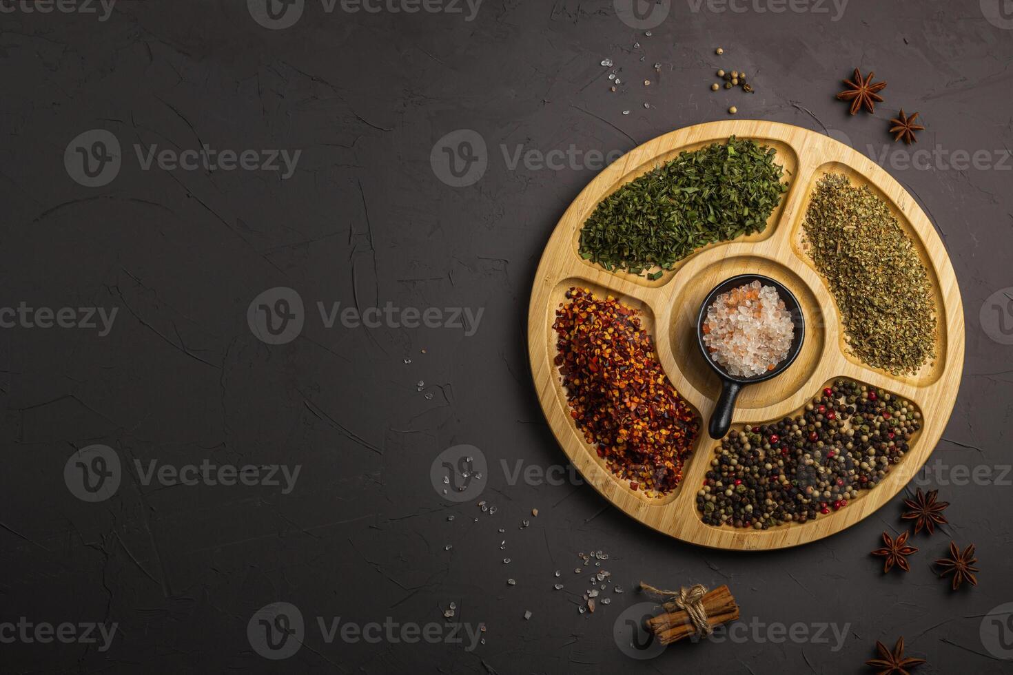 Mix spices on round wooden plate. Coriander seeds, ground red pepper, salt, black pepper, rosemary, turmeric, curry. Top view, close up, gray stone background. photo