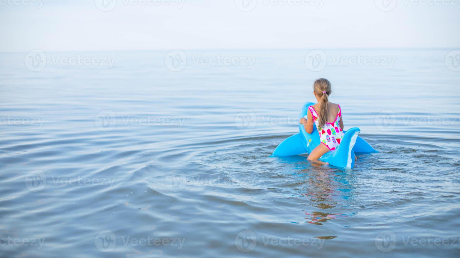 Happy girl of European appearance age of 7 swimming on an inflatable big shark toy in the sea.Family summer vocation concept. Copy space.Back view . photo