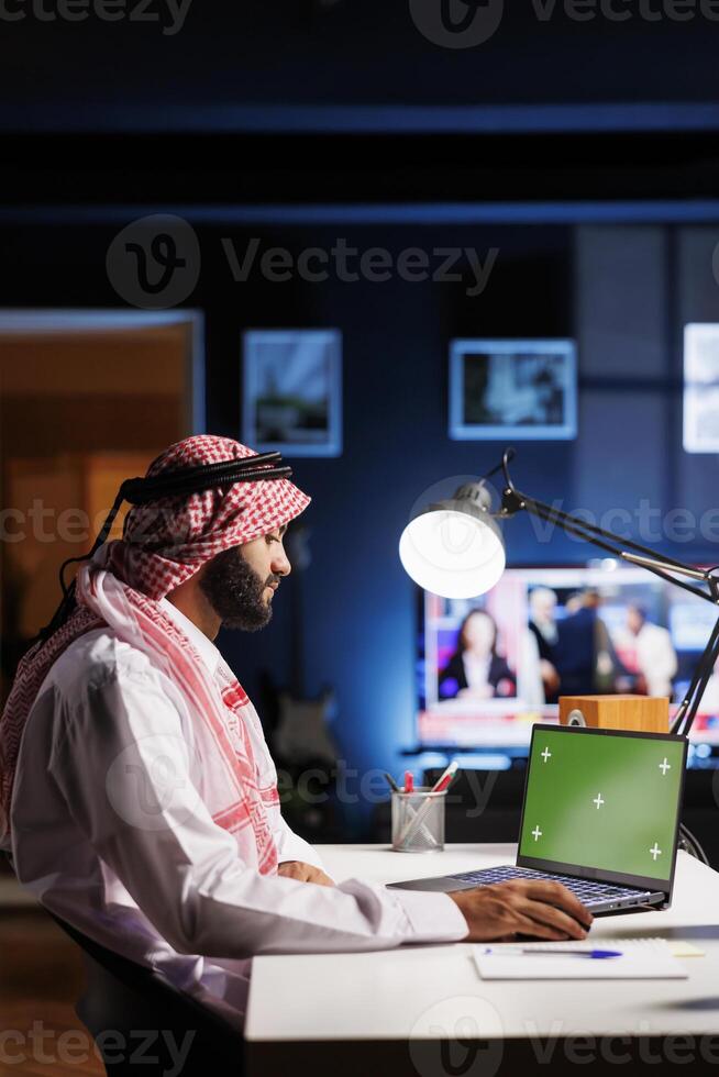 Arab male professional wearing traditional attire is seated at an office desk, using a laptop with an empty green screen. The image portrays a modern workspace for advertising or presentations. photo