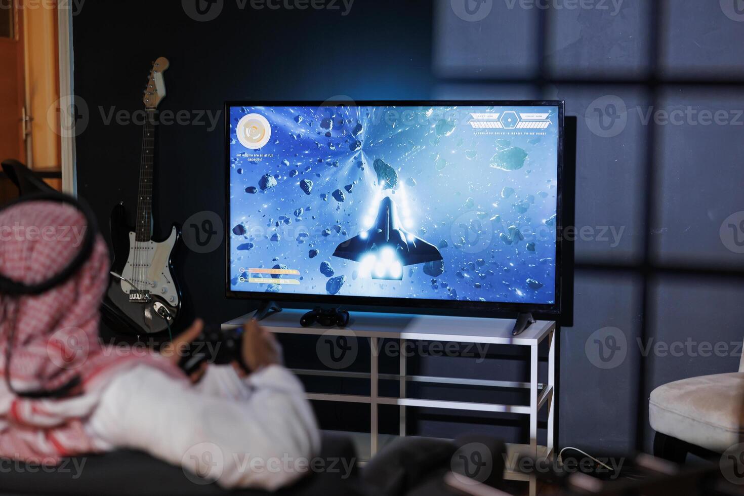 A Middle Eastern man immersed in the digital world with a wireless controller. Over-the-shoulder shot of a male Arab person seated in front of his television playing video games. photo