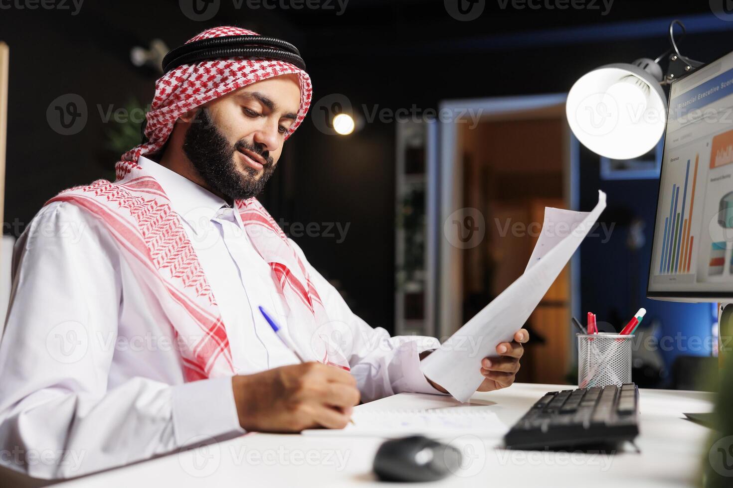 Islamic man working at his desk, surrounded by a desktop computer, documents, and paperwork, showcasing a busy corporate workspace. Side-view shot of Muslim guy writing down notes from research. photo