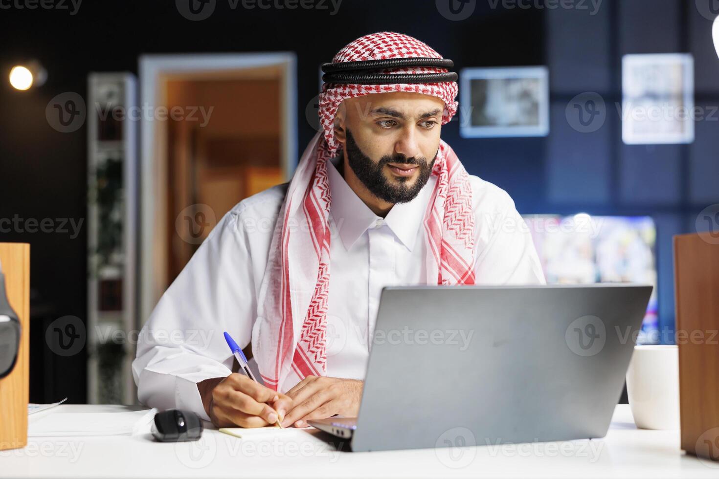 A focused Arab entrepreneur efficiently works on his laptop at a well-organized office desk. He types, browses the internet, and takes notes while surrounded by modern technology. photo