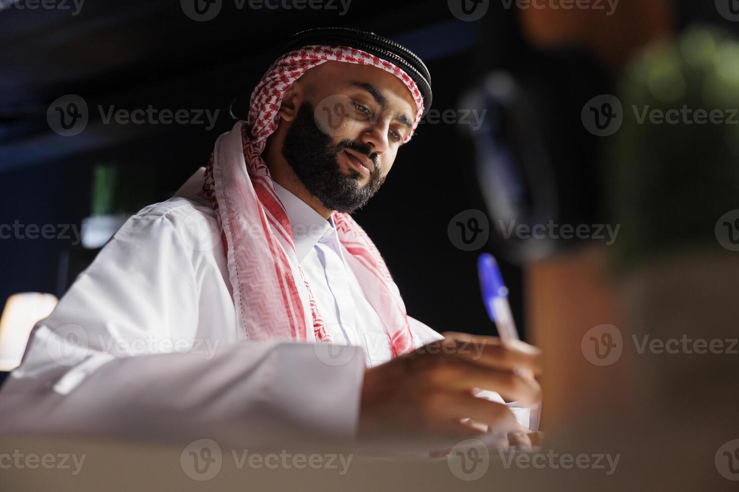 Detailed image showcasing focused Arab entrepreneur at the table writing on a notebook. Close-up shot of a Muslim man using a pen for taking research notes in his home office. photo