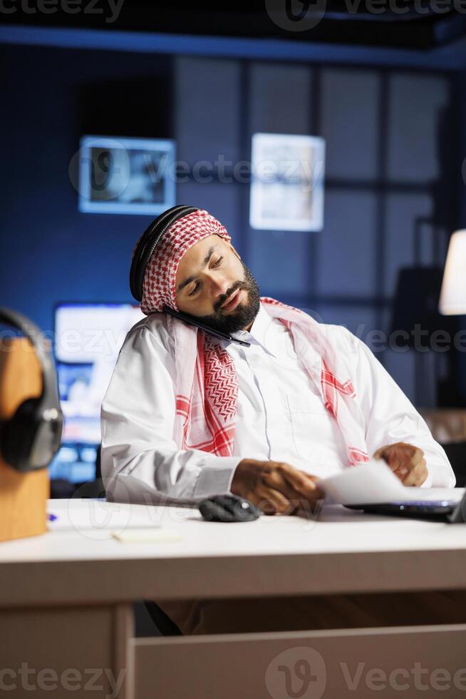 musulmán empresario en tradicional atuendo, Hablando en su Teléfono móvil y comprobación notas en un ordenador portátil. árabe empresario conversando con un compañero de trabajo y comparando investigación en un miniordenador. foto