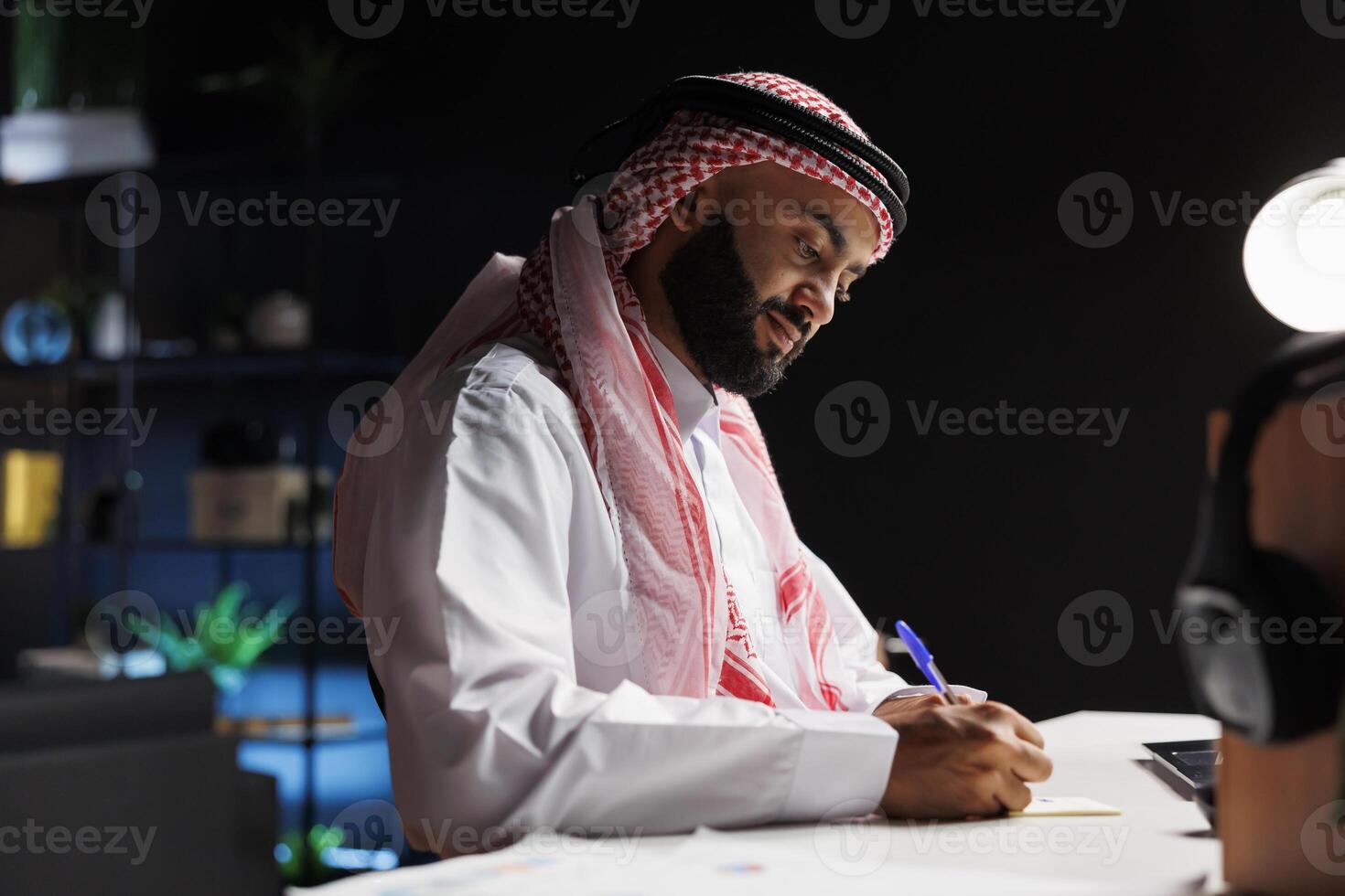 At his desk, a businessman from the Arab world skillfully browses the internet while focused on his work. He performs internet research while taking notes, demonstrating his expertise and commitment. photo