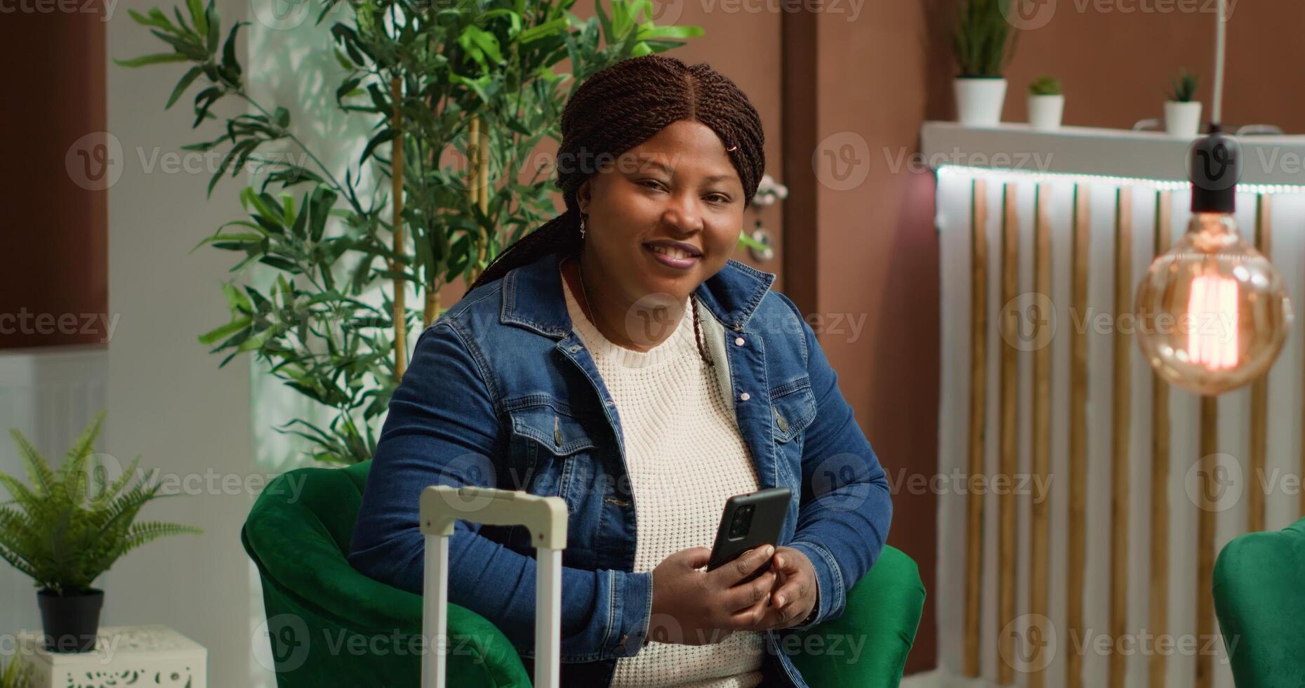 Woman visitor using phone in lounge area, checking social media messages while waiting for check in time. African american tourist on webpage arriving at five star hotel, travelling on holiday. photo
