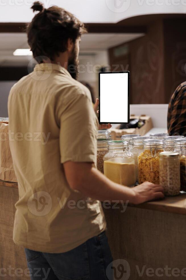 Client in zero waste shop using mockup tablet to videocall his girlfriend, buying her farm grown food. Man with isolated screen device phone calls partner over internet in local grocery store photo