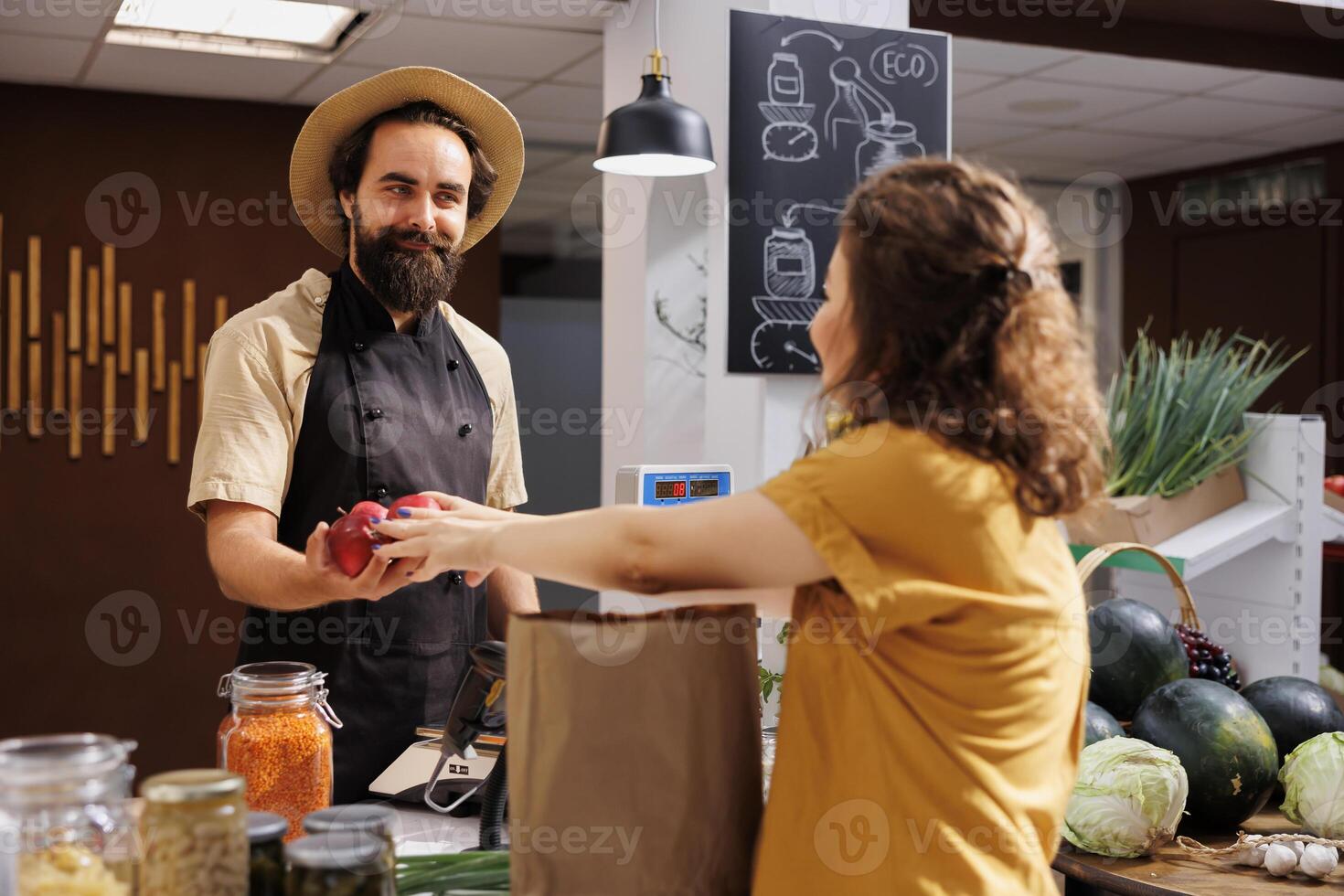 Woman at zero waste store checkout counter paying for pesticides free vegetables. Client in plastic free local grocery shop using decomposable paper bag for low carbon footprint photo