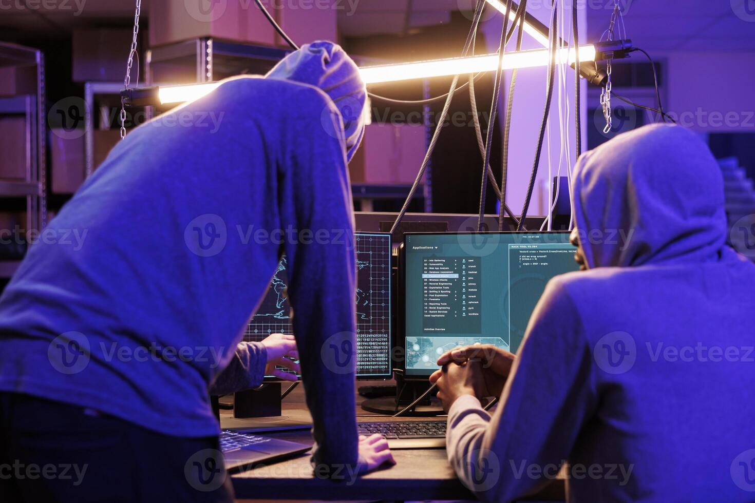 Two hackers running malicious software on computer screen to breach secure data. Criminals group wearing hoodies while breaking law and working together in abandoned warehouse photo