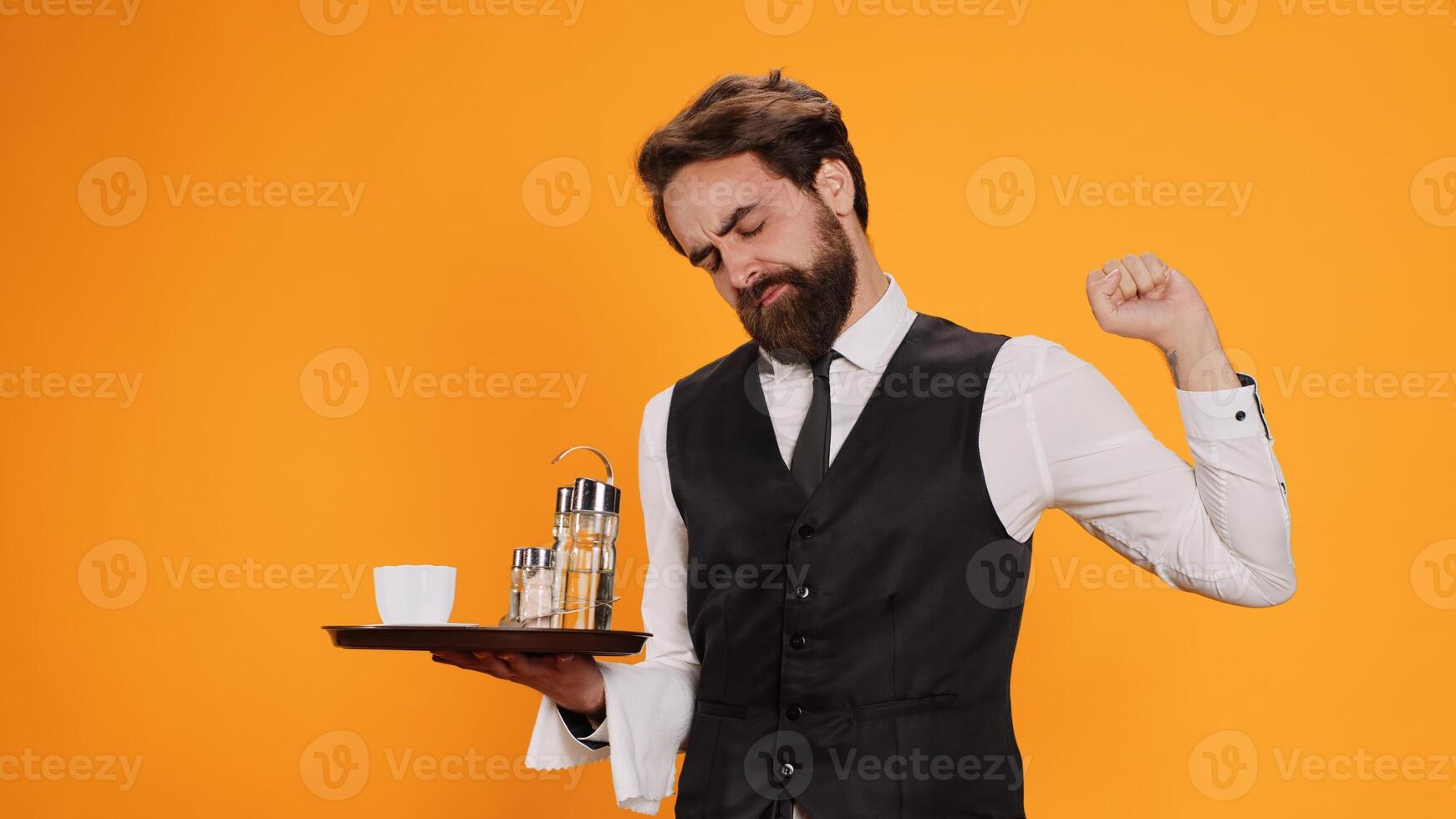 Overworked waiter feeling tired on camera, yawning and stretching on duty. Experienced employee in suit and tie feeling tired and almost falling asleep in studio, poses in yellow studio. photo