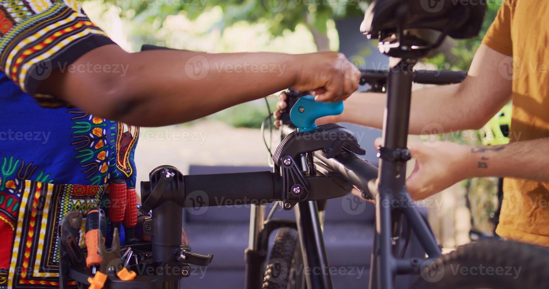 Healthy african american girlfriend assisting her caucasian boyfriend fix his bicycle outside. Active young woman fastening bike to repair-stand for athletic man to repair in home yard, closeup. photo