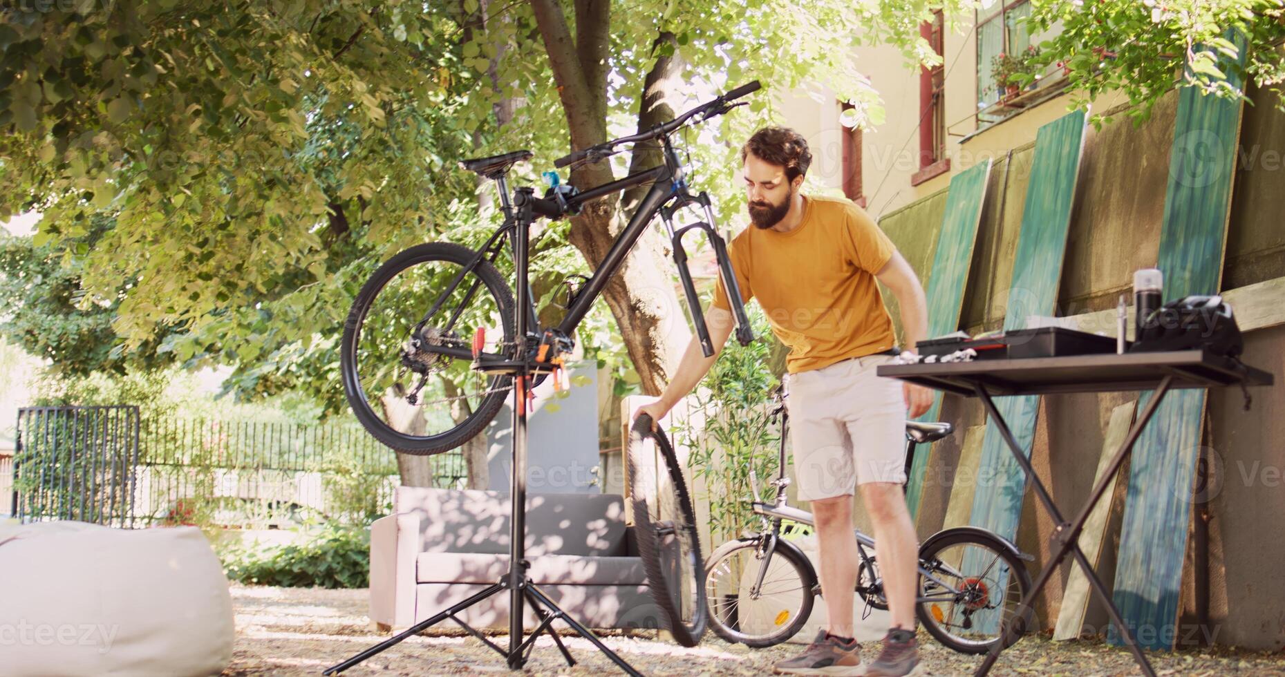 Athletic dedicated man fixing damaged bicycle with specialized equipment from toolbox in yard. Healthy caucasian male dismantling wheel and servicing bike front fork. Pull-back shot. photo