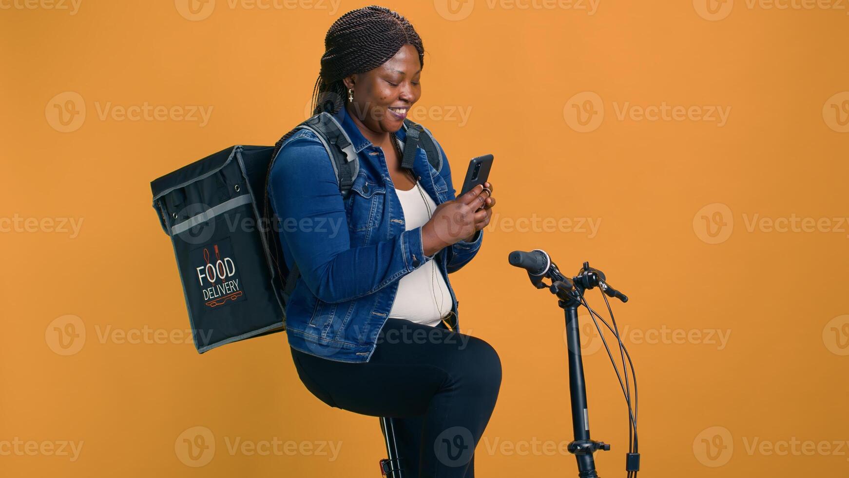 Smiling black woman texting coworkers with phone on bicycle before cycling for food delivery service job. African american lady checking mobile app for packages ready for delivering. photo